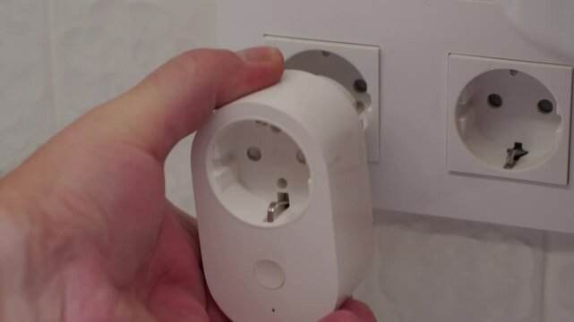 A white plastic smart plug, held by a hand, is being inserted into a wall power socket. The metal prongs connect with the outlet's contacts, producing a slight metallic click
