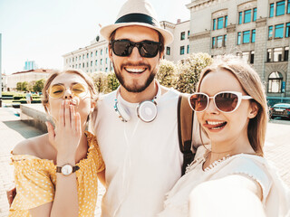 Group of young three stylish friends in the street.Man and two cute girls dressed in casual summer clothes.Smiling models having fun in sunglasses.Women and guy making photo selfie on smartphone
