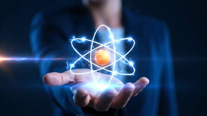 A person holds a glowing atom model in their hand, set against a dark blue backdrop. The hand, visible jacket sleeve, and lens flare highlight scientific innovation. 