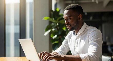 Happy african american man working on laptop in modern office, typing on keyboard. Business communication, remote work, or online learning concept.