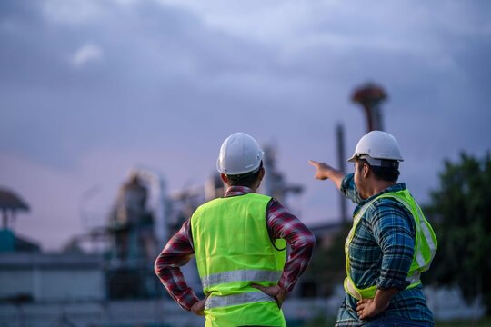 Engineers wearing safety helmets and reflective vests discussing project direction while one points toward the industrial plant in the distance.