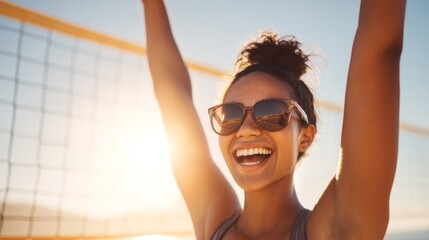 Joy radiates from a young woman with her arms raised in celebration, wearing polarized sunglasses. She enjoys an exciting moment on the beach volleyball court as the sun sets behind her