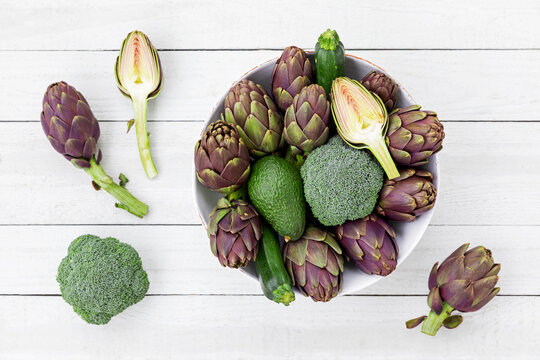 Fresh green artichoke broccoli zucchini and avocado in bowl on white table