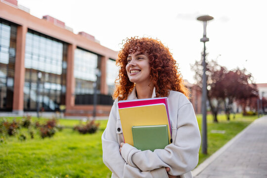 Student with curly red hair holding notebooks outdoors on a university campus