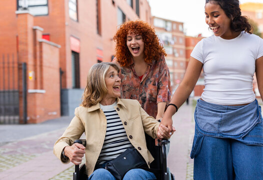 A mother in a wheelchair is assisted by two young women outdoors, showcasing togetherness and support.