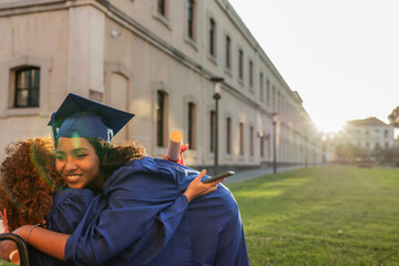 Two young women in graduation attire embracing on a university campus at sunset