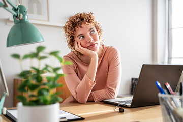 Thoughtful woman sitting at home office desk with laptop during daytime