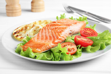 Piece of tasty grilled salmon, lemon slices and salad on white wooden table, closeup