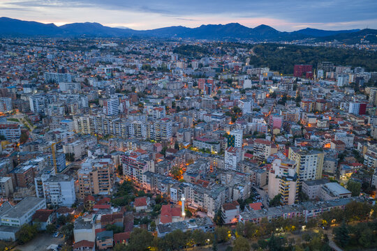 Dense urban neighbourhood of Tirana at blue hour, with layered residential blocks and Mount Dajti rising in the background before sunrise.