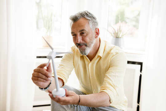 Mature man holding wind turbine model at home for sustainable energy
