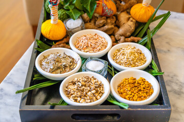 Thai herbal medicine ingredients in bowls on tray, featuring dried herbs, turmeric, ginger, salt, spa wellness concept.