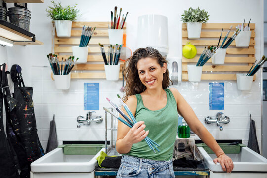 Smiling artist holding paintbrushes in art studio near washing station