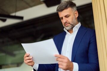 Focused Businessman In Blue Suit Reads Document At Modern Office Workspace During Important Review