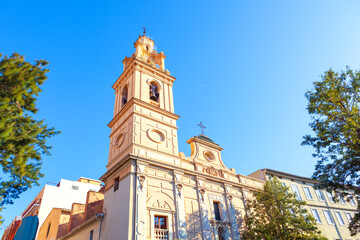 View of Church of El Salvador and Santa Monica in Valencia, distinctive bell tower rising against sky. Green trees frame sides of historic building, which showcases blend of architectural styles