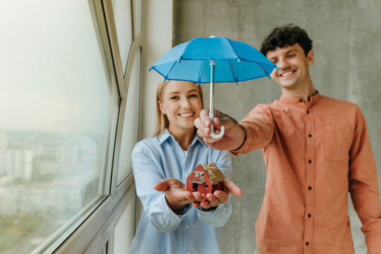 Couple protecting house model with umbrella indoors showing happiness