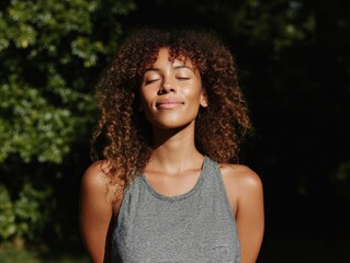 Woman breathing deeply in nature, curly hair glowing, wearing gray tank top. Peaceful moment shows calm and wellness in green forest.