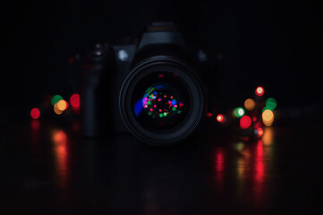 Close-up DSLR camera on dark background with festive bokeh lights reflected in the lens. Creative photography concept, low key, holiday ambiance.