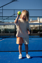 Blond young boy standing on vivid blue tennis court near black net, holding a green tennis ball in raised hand, dressed casually in beige t-shirt and orange shorts
