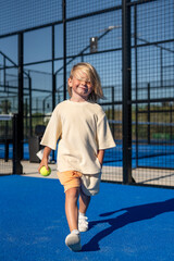 Smiling blond young boy walking on blue tennis court, holding a green tennis ball in one hand, dressed in beige oversized t-shirt, orange shorts, and white sneakers in sunlight