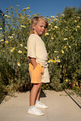 Blond young boy standing outdoors in front of blooming daisies, wearing casual beige oversized t-shirt, orange shorts, and white sneakers, hands in pockets, looking calmly under clear sunny sky