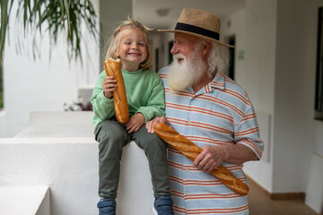 Elderly man with a long white beard and straw hat smiling while holding a baguette, as his young grandson in a green sweatshirt playfully takes a bite of his own bread