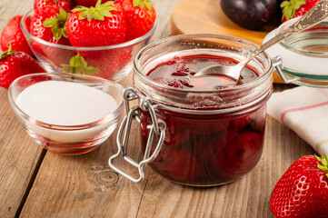 Jar of strawberry jam on a wooden table with fresh strawberries and a bowl of sugar in the background, creating a rustic homemade food and fruit concept ideal for culinary or recipe projects