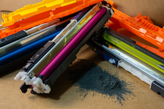 Close-up of colorful printer cartridges with spilled black toner powder on a cardboard background.