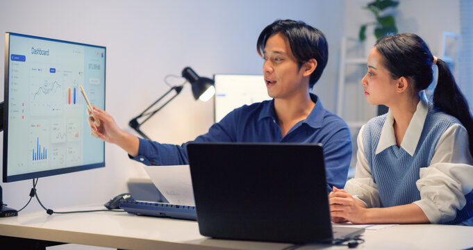 Asian male business leader explaining financial dashboard data on a monitor to a female colleague late at night. Conveys consulting, professional discussion, and dedicated teamwork.