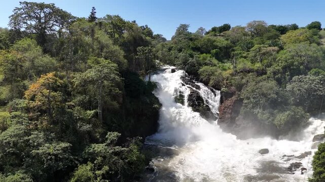 Lateral aerial footage of Binga Falls in Angola, showing the water plunging through thick jungle vegetation under bright tropical light and mist
