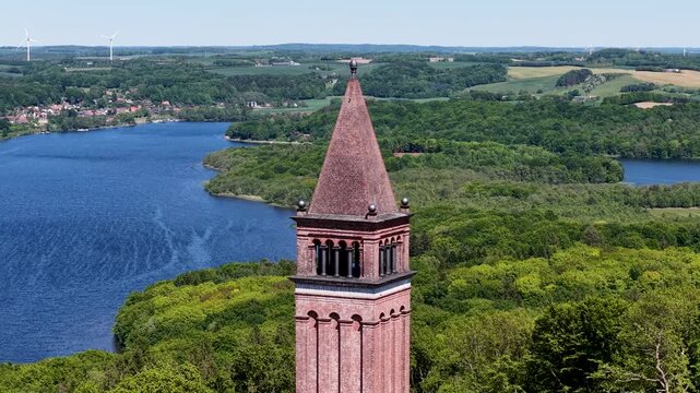 Drone footage capturing the iconic Himmelbjerget sky mountain tower rising above lush green forests and deep blue lake in Denmark, offering a scenic aerial view of the surrounding landscape.