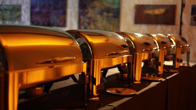 A row of golden chafing dishes on a banquet table, reflecting warm ambient lighting, set against a backdrop of abstract wall art. Event catering concept