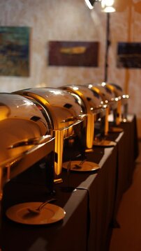 A row of golden chafing dishes on a banquet table, reflecting warm ambient lighting, set against a backdrop of abstract wall art. Event catering concept