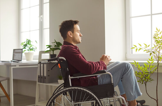 Handicapped young man posing in wheelchair and looking through window, has problems with mobility after accident, feels lonely as being at home alone. People and health problems concept