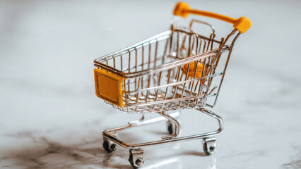 A miniature chrome shopping cart with yellow accents sits on a light marble surface, symbolizing online shopping, retail, and consumerism.