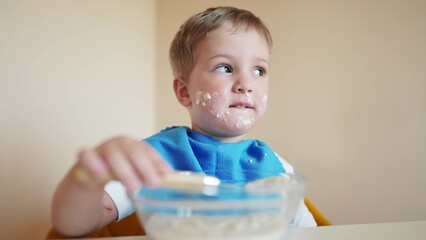 baby eats porridge. grimy a blond kid boy soiled his face eats himself porridge with a spoon. happy family kindergarten kid dream concept. kid eats in kindergarten lifestyle himself dirty face