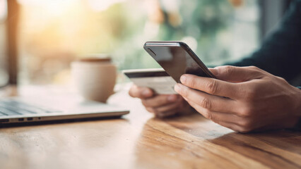 Hands holding phone and credit card for online payment or shopping, near a laptop and coffee cup on a wooden desk.
