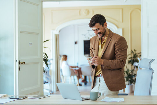 Businessman Smiles While Using Phone in Modern Office With Laptop and Coffee Mug - Powered by Adobe