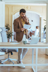 Businessman on Phone Reading Paperwork in Modern Office Desk Setup with Laptop and Coffee Workspace