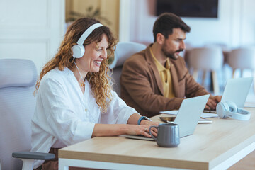 Smiling Woman With Headphones Working At Shared Desk While Man Focuses On Laptop In Modern Office