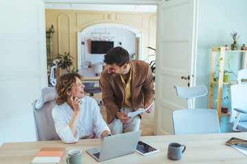 Friendly Office Collaboration Between Colleagues Laughing While Reviewing Documents and Laptop in Open Plan Workspace