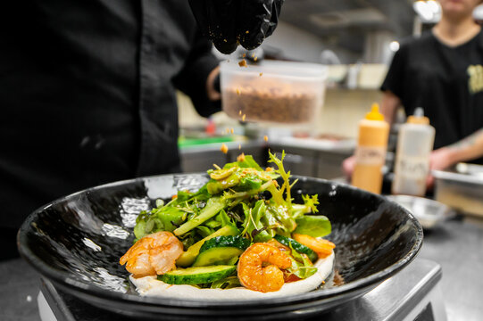 A close-up shot of a professional chef wearing black gloves, adding garnish to a fresh and vibrant shrimp salad served in a black bowl within a commercial kitchen setting.
