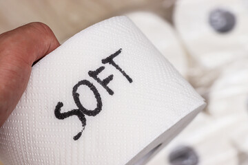 A hand holding a roll of toilet paper labeled with the word SOFT. The focus is on the texture and...