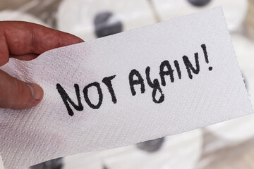 A hand holds a white paper towel displaying the handwritten message NOT AGAIN! emphasizing a sense of frustration. The background includes rolls of toilet paper.