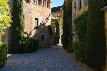 Charming cobbled street in Peratallada’s historic center, flanked by cypress trees, green bushes, and rustic stone buildings in warm light.