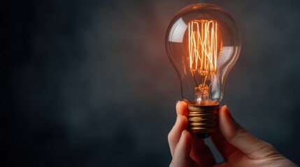 A hand holding a glowing incandescent light bulb with visible filament brightening the dark background,