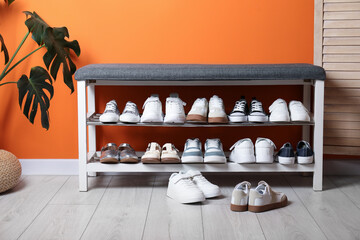 Different kid's shoes, storage bench and houseplant near orange wall indoors