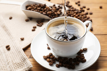 Pouring aromatic coffee into cup and beans at wooden table, closeup