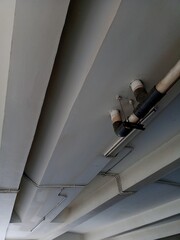 A low-angle, abstract view of a ceiling or underside of an overpass, showing exposed support beams and plumbing pipes (white and black) in a grey industrial space.