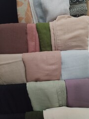 A grid-like pattern of neatly folded fabrics or scarves stacked vertically and horizontally, featuring a diverse palette of muted colors including pink, mauve, beige, olive, and navy.