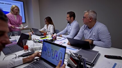 Group of diverse business professionals collaborates during a corporate meeting, analyzing data and discussing strategy. A female presenter distributes documents while colleagues work on laptops. - Powered by Adobe